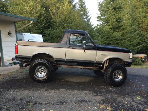 1989 Ford Bronco - A lifted two-tone Ford Bronco is parked on a gravel driveway next to a house and trailer.
