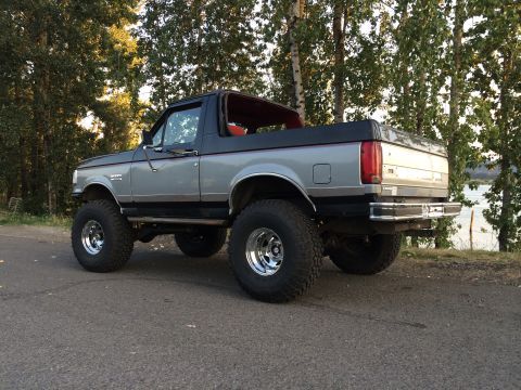 1989 Ford Bronco - A lifted 1989 Ford Bronco is parked on the side of a road with trees and water in the background.