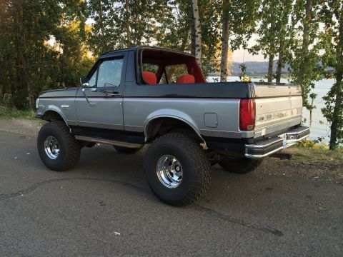 1989 Ford Bronco - A lifted, two-tone Ford Bronco with an orange interior is parked on a road near trees and a body of water.