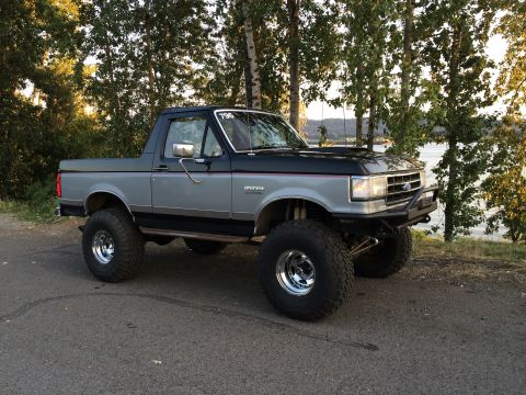 1989 Ford Bronco - A lifted 1989 Ford Bronco with a two-tone paint job sits parked outdoors near a body of water.