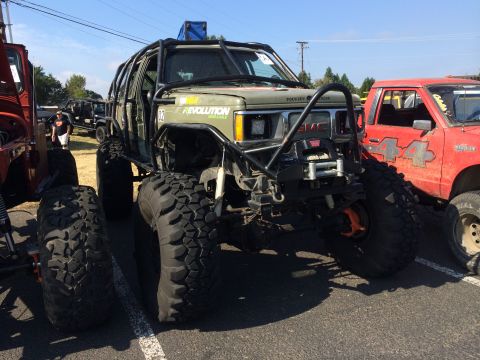 1989 Ford Bronco - A heavily modified GMC SUV with a roll cage and massive off-road tires is parked next to other vehicles at an outdoor event.