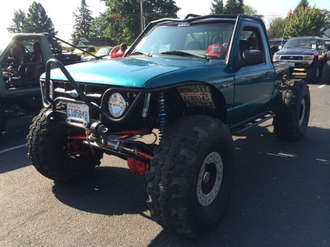 1989 Ford Bronco - A modified teal pickup truck with oversized tires and a roll cage is displayed at an outdoor event.