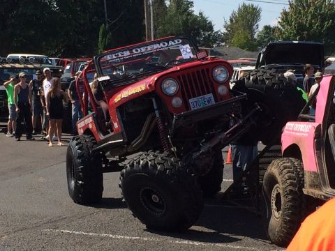 1989 Ford Bronco - A heavily modified red jeep with oversized tires is displayed at an off-road event, with other vehicles and spectators in the background.