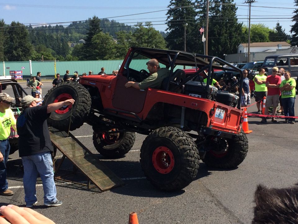 Red Jeep with large tires on a ramp, driver inside, people watching
