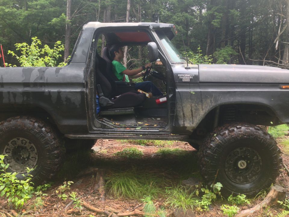Side view of a 1979 Ford Bronco with the door open, a person inside driving on a wooded trail