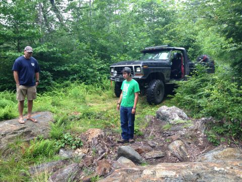 1979 Ford Bronco - A black Ford Bronco navigates a wooded, rocky terrain with two onlookers present.