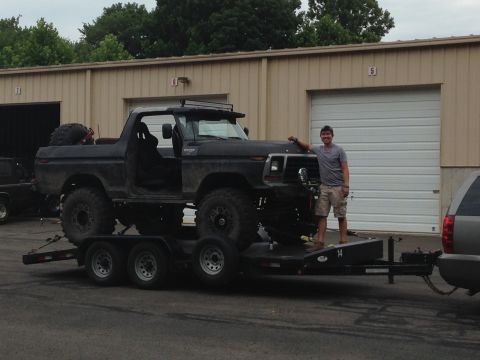1979 Ford Bronco - A man stands beside a heavily modified Ford Bronco loaded onto a trailer in front of a garage.