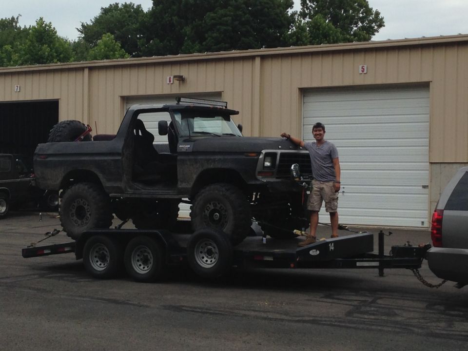 Guy next to a lifted black Ford Bronco on a trailer, doors removed, with a spare tire in the bed