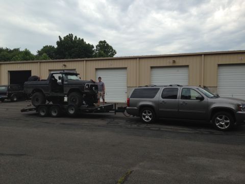 1979 Ford Bronco - A black Ford Bronco is loaded onto a trailer, towed by a grey SUV in front of a row of garage bays.