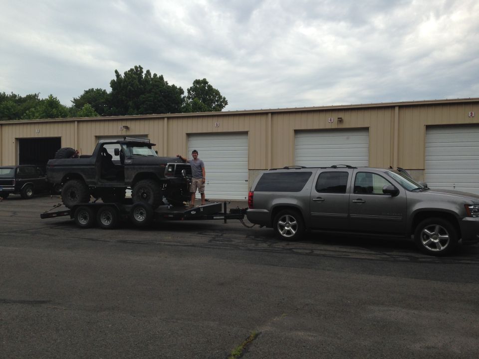 Black 1978 Ford Bronco on a triple-axle trailer, towed by a grey Chevrolet Suburban