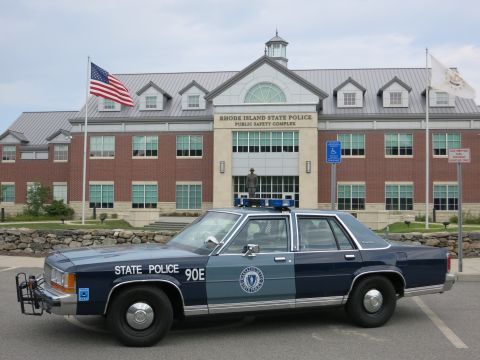 1991 Ford LTD Crown Victoria - two-tone blue State Police sedan marked 90E parks before the brick Rhode Island State Police Public Safety Complex under a U.S. flag.