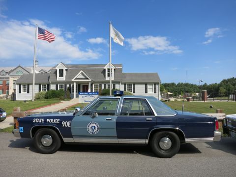1991 Ford LTD Crown Victoria - two-tone blue State Police sedan marked 90E sits before a gray-shingled museum building flying U.S. and state flags.