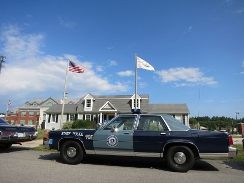 1991 Ford LTD Crown Victoria - two-tone blue State Police 90E sedan is parked before a shingled museum building under U.S. and state flags on a clear day.
