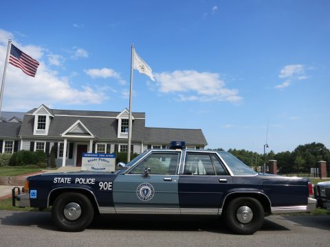 1991 Ford LTD Crown Victoria - black-and-white police cruiser with roof lights and a silver sedan sit on a roadway near a flagpole and shingled buildings.