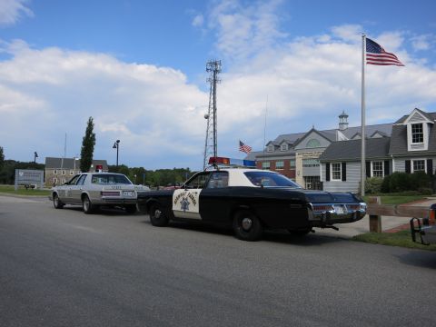 1991 Ford LTD Crown Victoria - white full-size sedan with a front push bar and a dark sedan are parked in a lot ringed by trees under a clear blue sky.