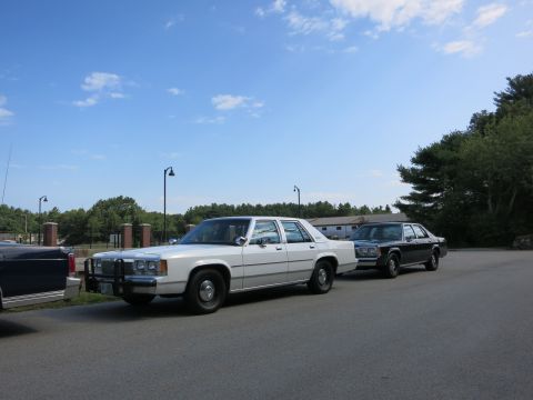 1991 Ford LTD Crown Victoria - line of vintage police cars including a white cruiser is parked along a roadside near a radio tower and shingled buildings.