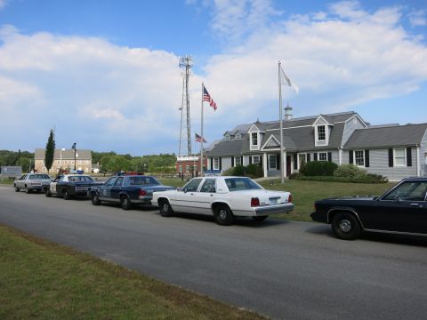 1991 Ford LTD Crown Victoria - row of vintage police sedans including a white cruiser lines a roadside before gray-shingled buildings and a radio tower.