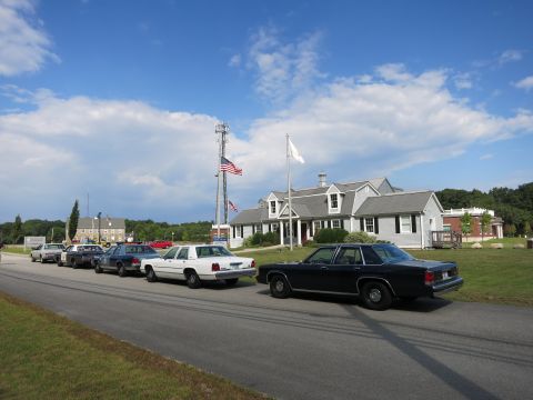1991 Ford LTD Crown Victoria - line of police cars is parked along a roadside near a tall radio tower, flagpoles, and gray-shingled buildings.