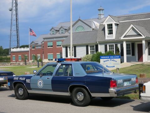 1991 Ford LTD Crown Victoria - two-tone blue State Police 90E sedan with roof lights parks before the Rhode Island State Police Museum sign and buildings.