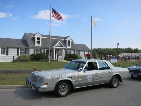 1991 Ford LTD Crown Victoria - silver sedan lettered Rhode Island State Police with a front push bar parks before a shingled building under a U.S. flag.