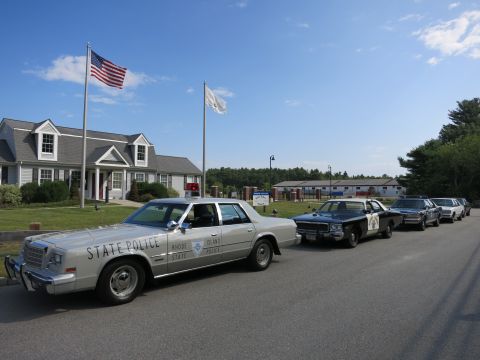 1991 Ford LTD Crown Victoria - silver Rhode Island State Police sedan leads a row of black-and-white cruisers parked along a roadside near flagpoles.
