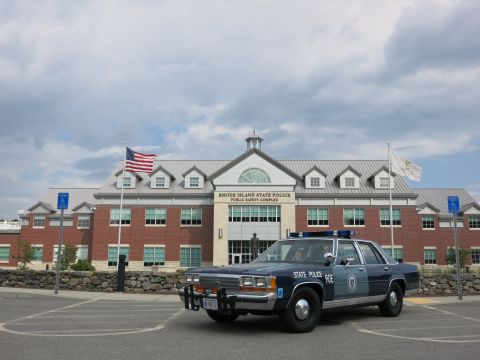 1991 Ford LTD Crown Victoria - Front three-quarter view of a blue State Police 90E sedan with a chrome push bar, parked outside the Rhode Island State Police brick buildi…