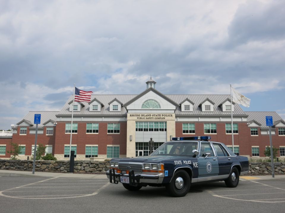 Front three-quarter view of blue State Police 90E sedan with push bar at brick complex