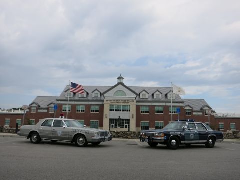 1991 Ford LTD Crown Victoria - silver State Police sedan and a blue State Police sedan parked apart in front of the Rhode Island State Police complex under a cloudy sky.