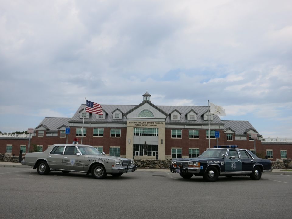 Silver and blue State Police sedans parked apart under cloudy sky at brick complex