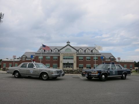1991 Ford LTD Crown Victoria - silver State Police sedan and a blue State Police sedan parked side by side facing forward in front of the Rhode Island State Police comple…