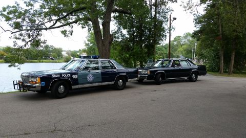 1991 Ford LTD Crown Victoria - Two dark blue State Police sedans, one marked 90E, parked on a tree-lined road beside a body of water.