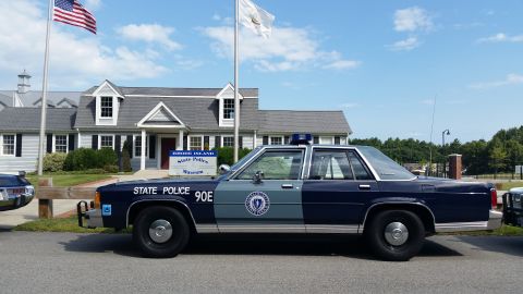 1991 Ford LTD Crown Victoria - blue State Police 90E sedan parked in profile near flagpoles outside the Rhode Island State Police Museum.