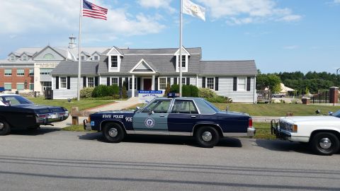 1991 Ford LTD Crown Victoria - blue State Police 90E sedan parked among a line of older vehicles in front of the white Rhode Island State Police Museum.