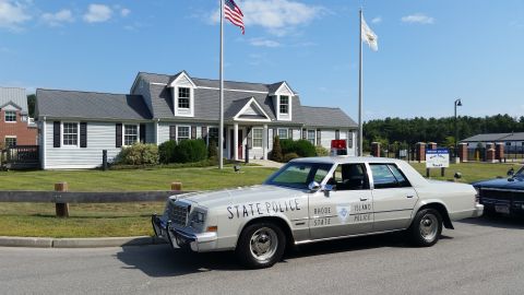 1991 Ford LTD Crown Victoria - cream-colored Rhode Island State Police sedan parked in profile near flagpoles in front of a white-sided building.