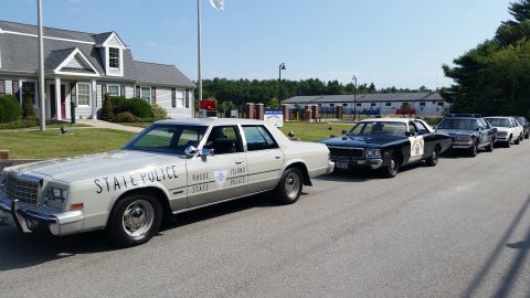 1991 Ford LTD Crown Victoria - cream Rhode Island State Police sedan leading a line of older police cars, including a black-and-white cruiser, outside a white building.
