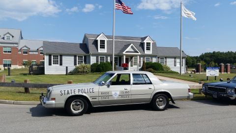 1991 Ford LTD Crown Victoria - cream-colored Rhode Island State Police sedan with a red roof light parked in profile by a flagpole outside a white building.