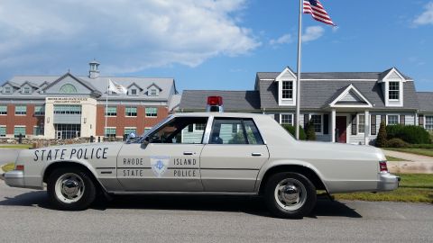 1991 Ford LTD Crown Victoria - cream Rhode Island State Police sedan parked in profile under an American flag between a brick complex and a white building.