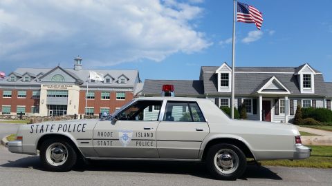 1991 Ford LTD Crown Victoria - cream Rhode Island State Police sedan with a red roof light parked in profile under an American flag near brick and white buildings.