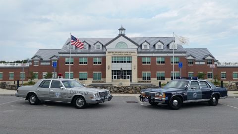 1991 Ford LTD Crown Victoria - A silver-gray Dodge Diplomat lettered 'RHODE ISLAND STATE POLICE' with a single red beacon and push bumper parked nose-to-nose-style alongs…