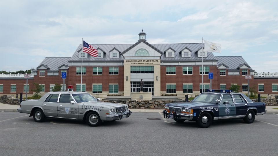 Silver Dodge and blue Ford state police cruisers parked outside a brick headquarters