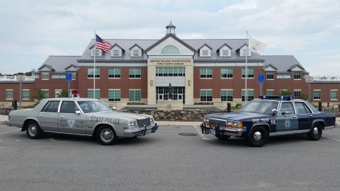 1991 Ford LTD Crown Victoria - Two restored police sedans nose-out in front of the brick Rhode Island State Police Public Safety Complex: a silver Chrysler-corporation cr…
