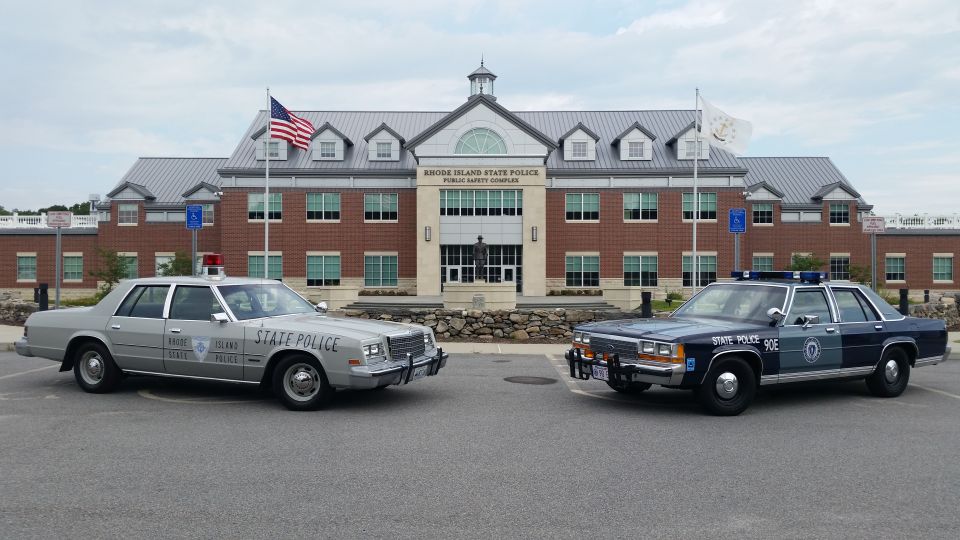Silver RI State Police sedan and blue Ford Crown Victoria cruiser parked at RISP headquarters