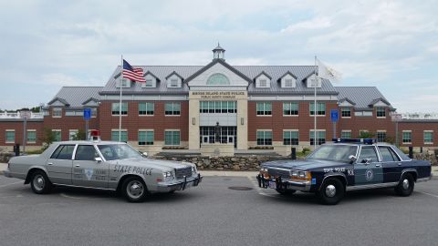 1991 Ford LTD Crown Victoria - Two restored cruisers parked side by side in front of the brick Rhode Island State Police Public Safety Complex: a silver-gray Dodge Diplom…