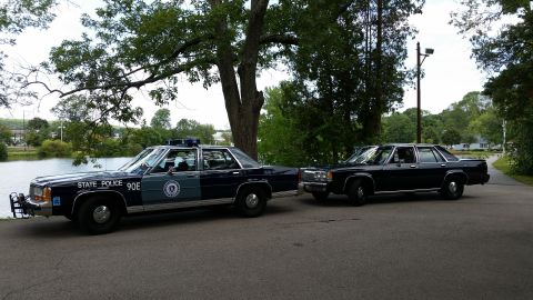 1991 Ford LTD Crown Victoria - Two dark blue Ford LTD Crown Victoria sedans parked side by side on pavement beside a pond and shade trees. The nearer car is a marked crui…