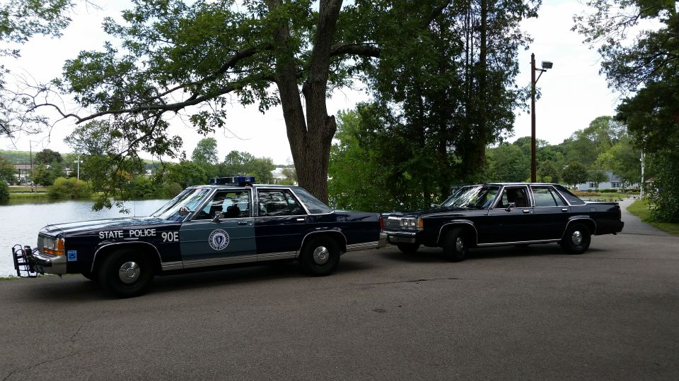 Two dark blue Ford Crown Victoria police sedans parked by a pond, marked and unmarked