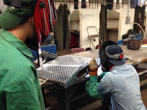 1979 Ford Bronco - Two individuals in welding masks work on a diamond plate surface in a vocational training setting.