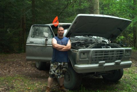 1979 Ford Bronco - A man in a sleeveless blue shirt and camo shorts stands with arms crossed in front of a dust-covered late-1970s Ford Bronco in the woods, h…