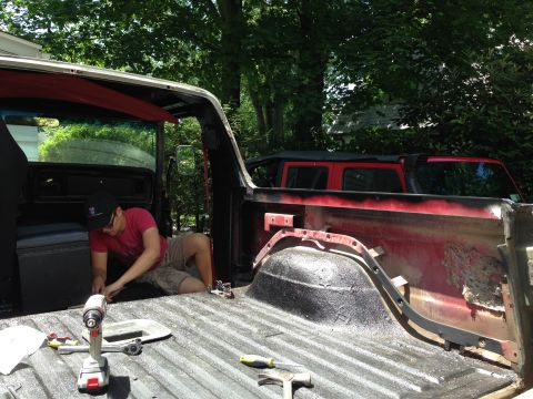 1979 Ford Bronco - A person works on the bed of a red pickup truck, with another vehicle visible in the background.