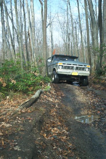 1979 Ford Bronco - Front three-quarter view of a lifted 1970s Ford 4x4 with a black body and white grille surround and lower panels, climbing a rutted muddy t…
