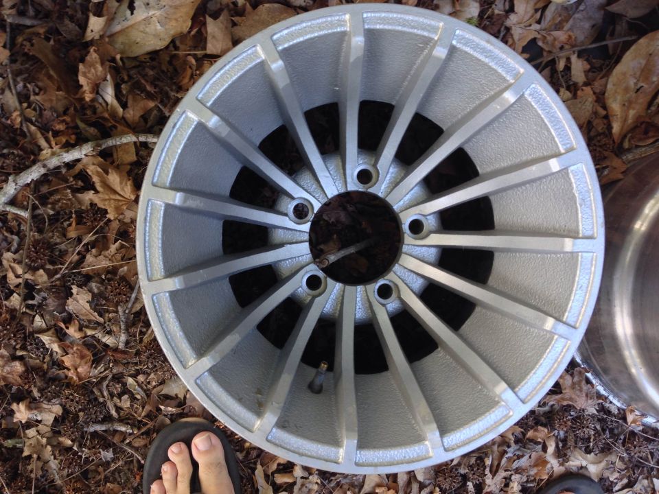 Close-up of a silver alloy car wheel with 15 spokes, resting on dry leaves. A person's foot in a sandal is visible at the bottom left